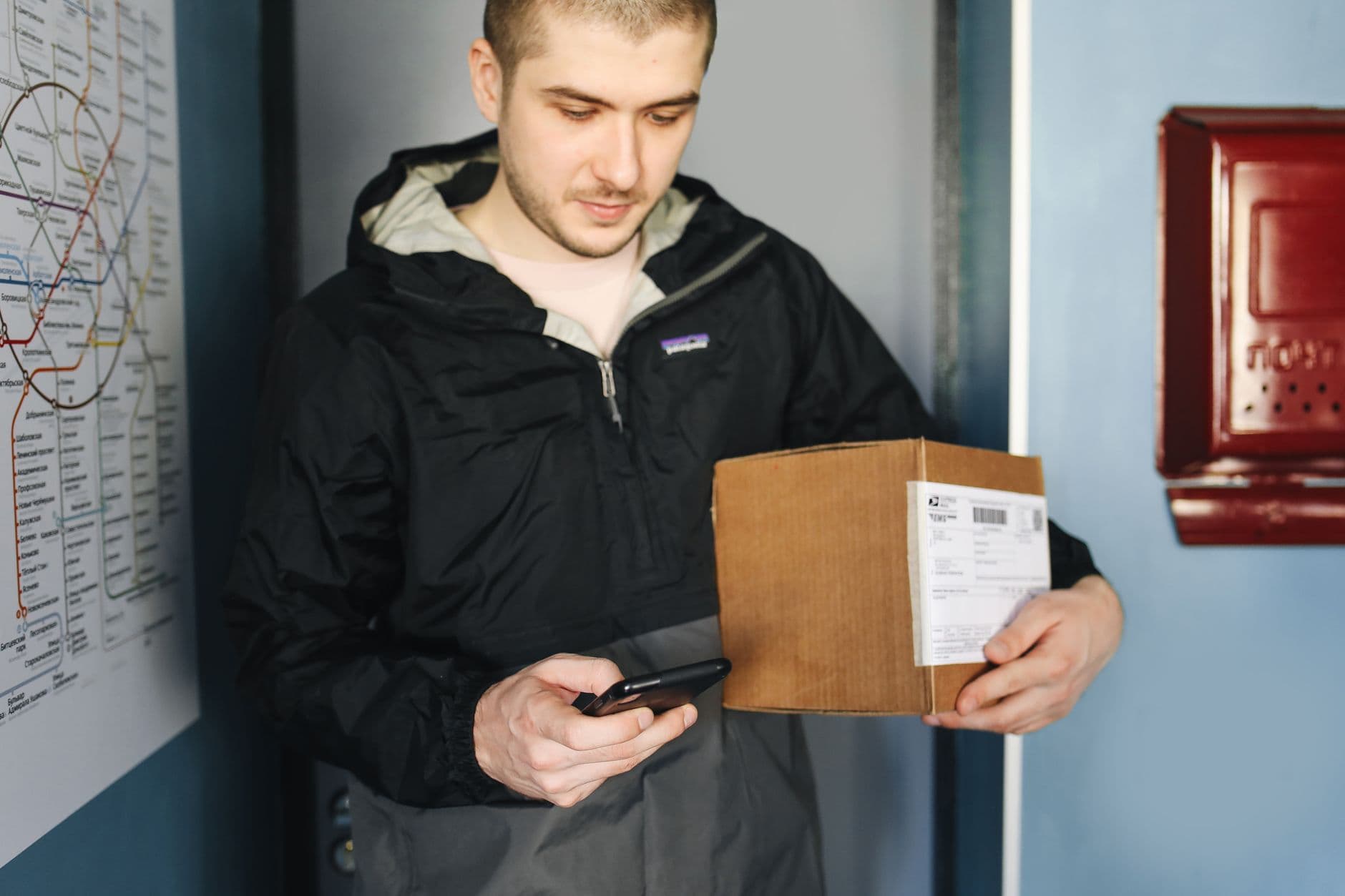 Stock photo of a courier in a rain jacket checking a phone and holding a labeled cardboard parcel beside a transit map
